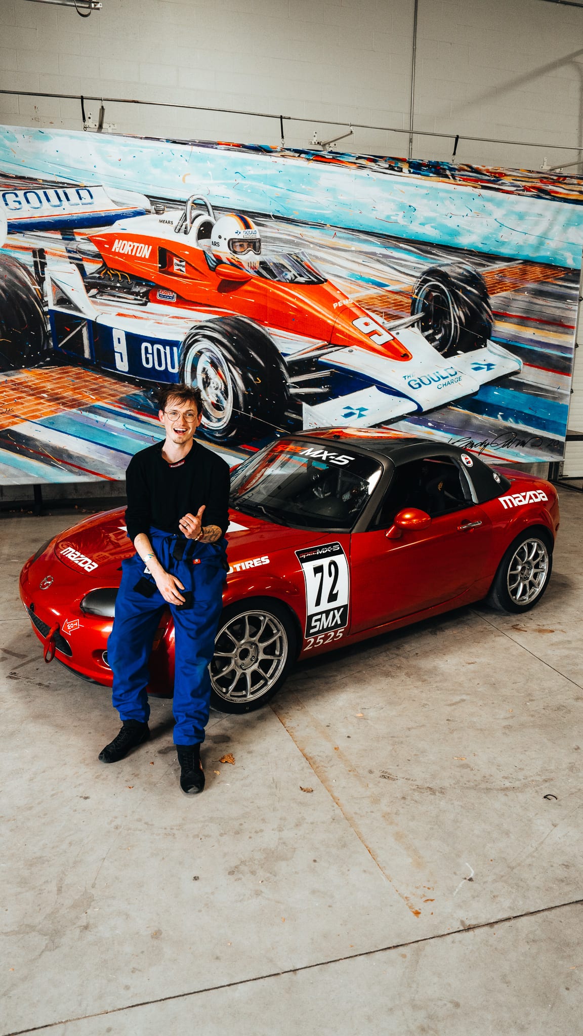 AlexMartini leaning against a red Spec MX-5 race car in front of a motorsport mural